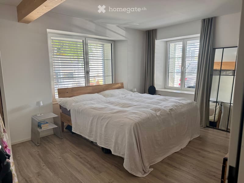 Modern bedroom with a double bed, white bedding, wooden nightstand, and a large mirror. The room features wooden flooring and large windows with blinds.