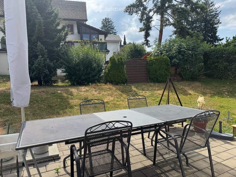 Garden with a patio area featuring a metal table and four chairs, surrounded by grass and trees, with a house visible in the background.