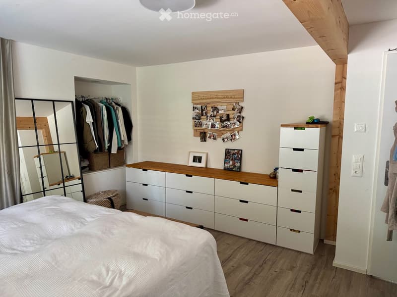 Modern bedroom with a large bed, white wardrobe, mirrored closet, and a wooden dresser with picture frames and baskets.