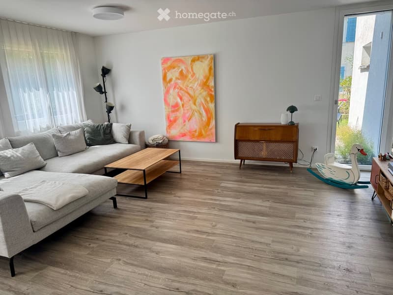 Modern living room with light wooden flooring, grey sectional sofa, wooden coffee table, side table, floor lamp, and a colorful abstract painting on the wall.