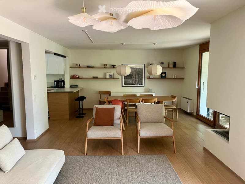 Open-plan living and dining area with light wood flooring, white walls, and modern decor. Features a sofa, armchairs, dining table with chairs, pendant lights, and a kitchen counter with appliances.
