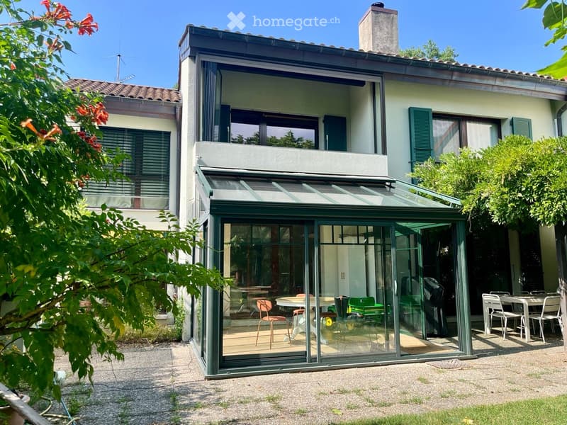 A modern house with a large glass-enclosed patio area, featuring outdoor furniture including tables and chairs, surrounded by greenery and a clear blue sky.