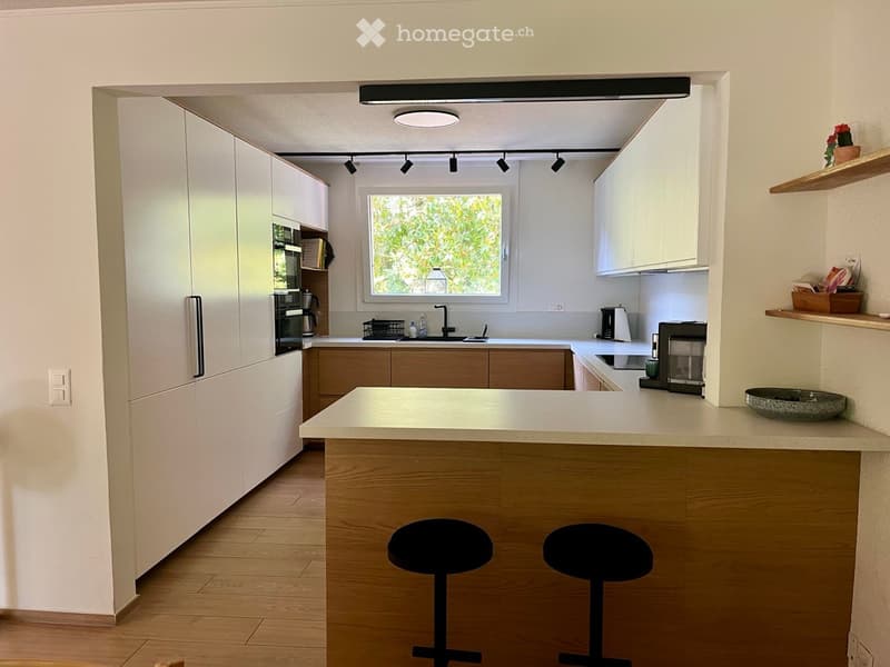 Modern kitchen with white cabinets, wooden countertops, and built-in appliances. Features a central island with two stools, a window above the sink, and various kitchen appliances including a refrigerator, oven, and coffee maker.