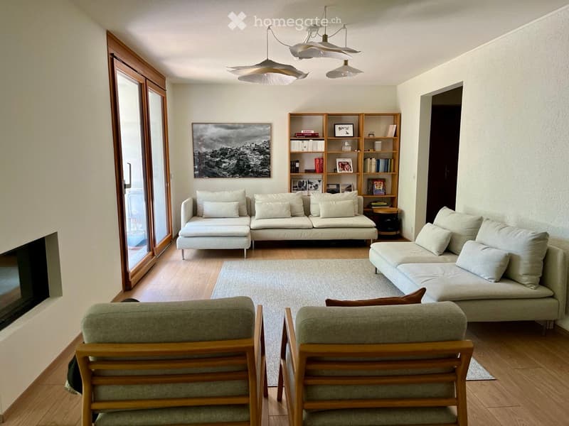 Modern living room with light-colored furniture, including a sectional sofa, two armchairs, a bookshelf, and a coffee table. The room features wooden flooring, large windows, and a fireplace.