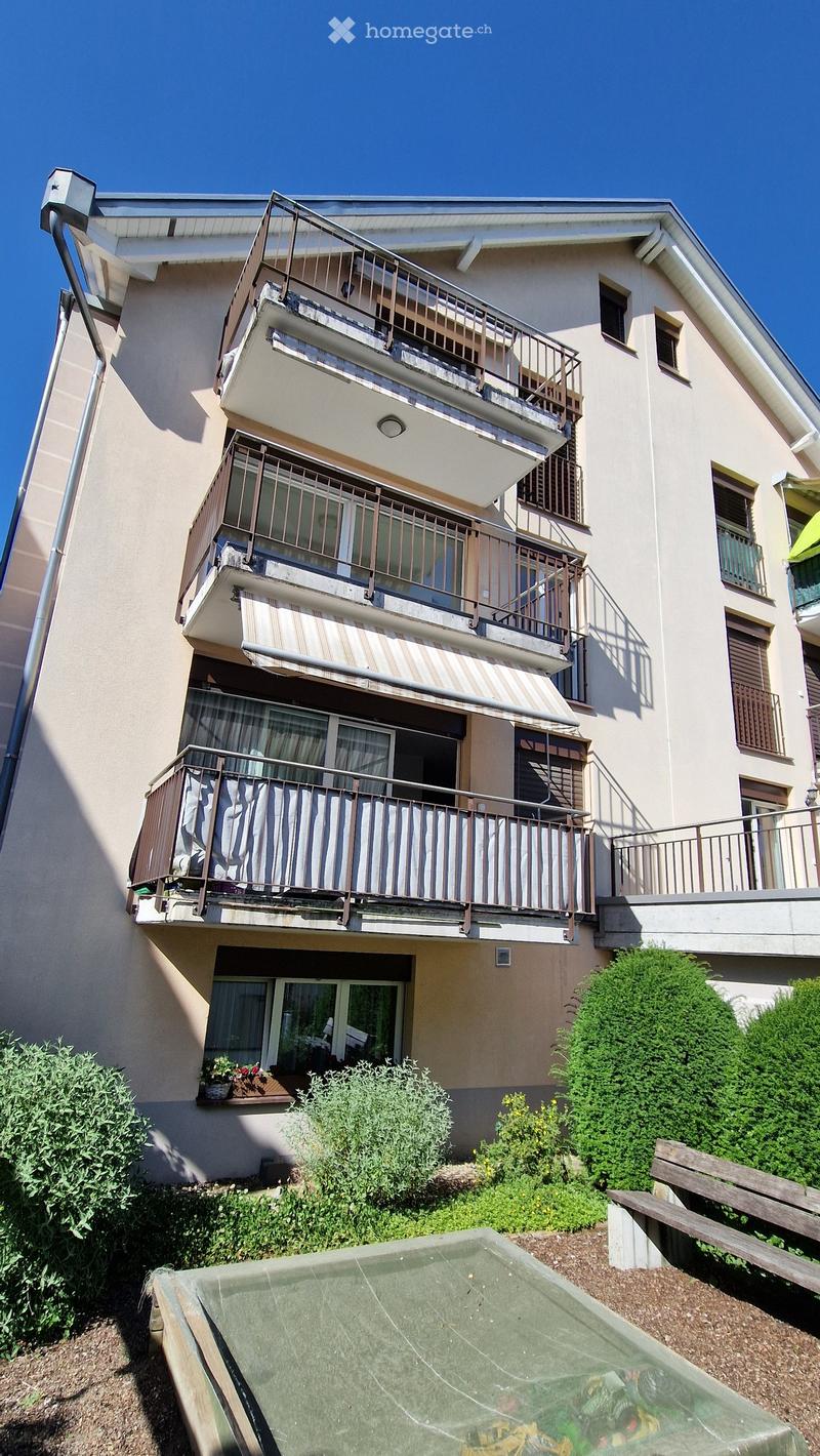 Three-story residential building with multiple balconies, beige facade, and small garden area in front.
