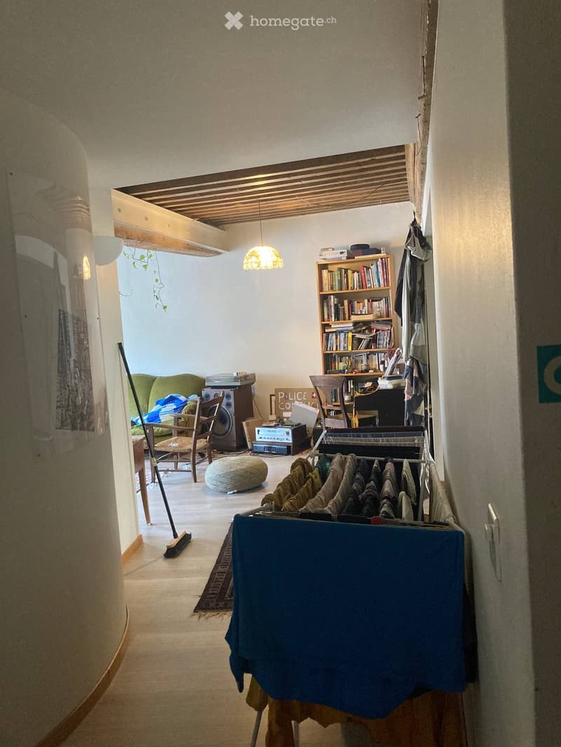 A cozy living space with a sofa, a wooden chair, a bookshelf filled with books, a small table, a round stool, a hanging lamp, a speaker, and a clothing rack with folded clothes.