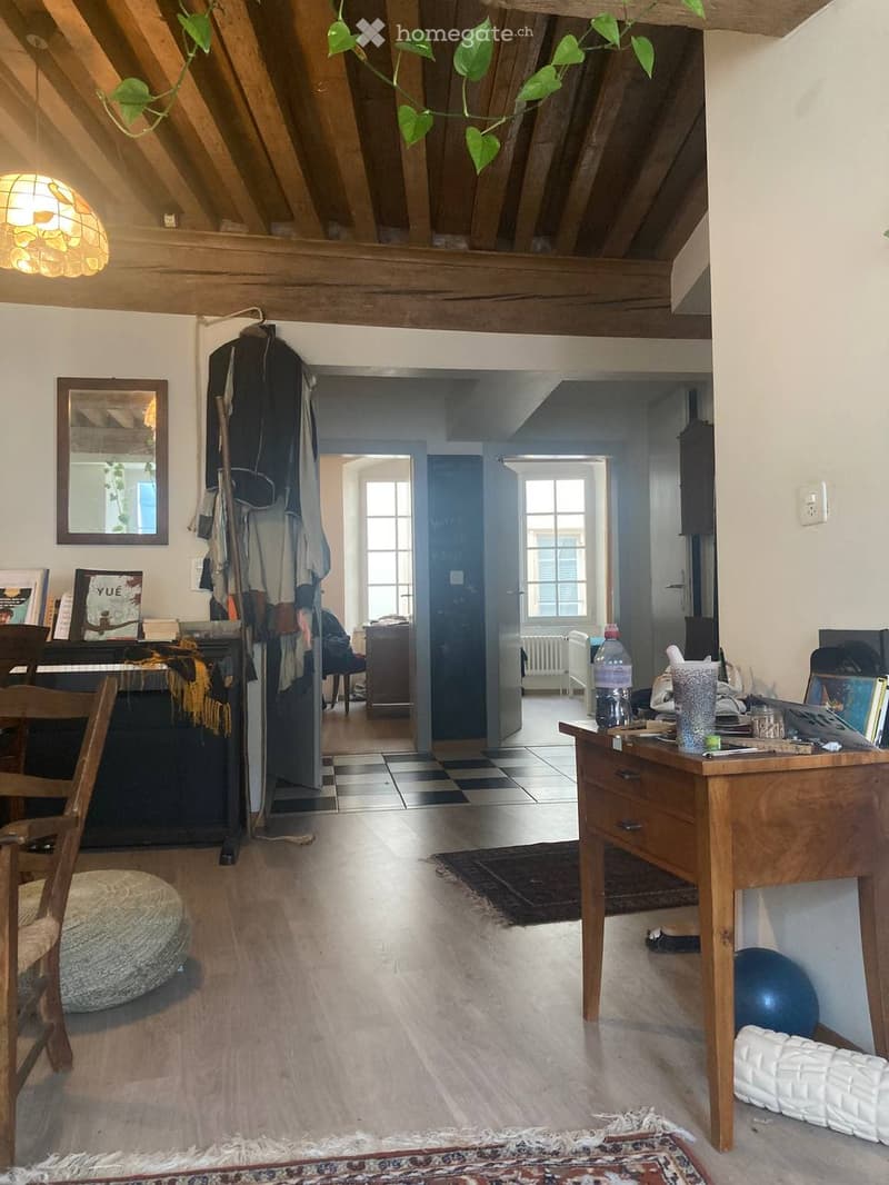 A cozy, well-lit room with wooden beams on the ceiling, a large window, and a mix of modern and traditional furniture including a chair, a round stool, a wooden table, a piano, and a ball on the floor.