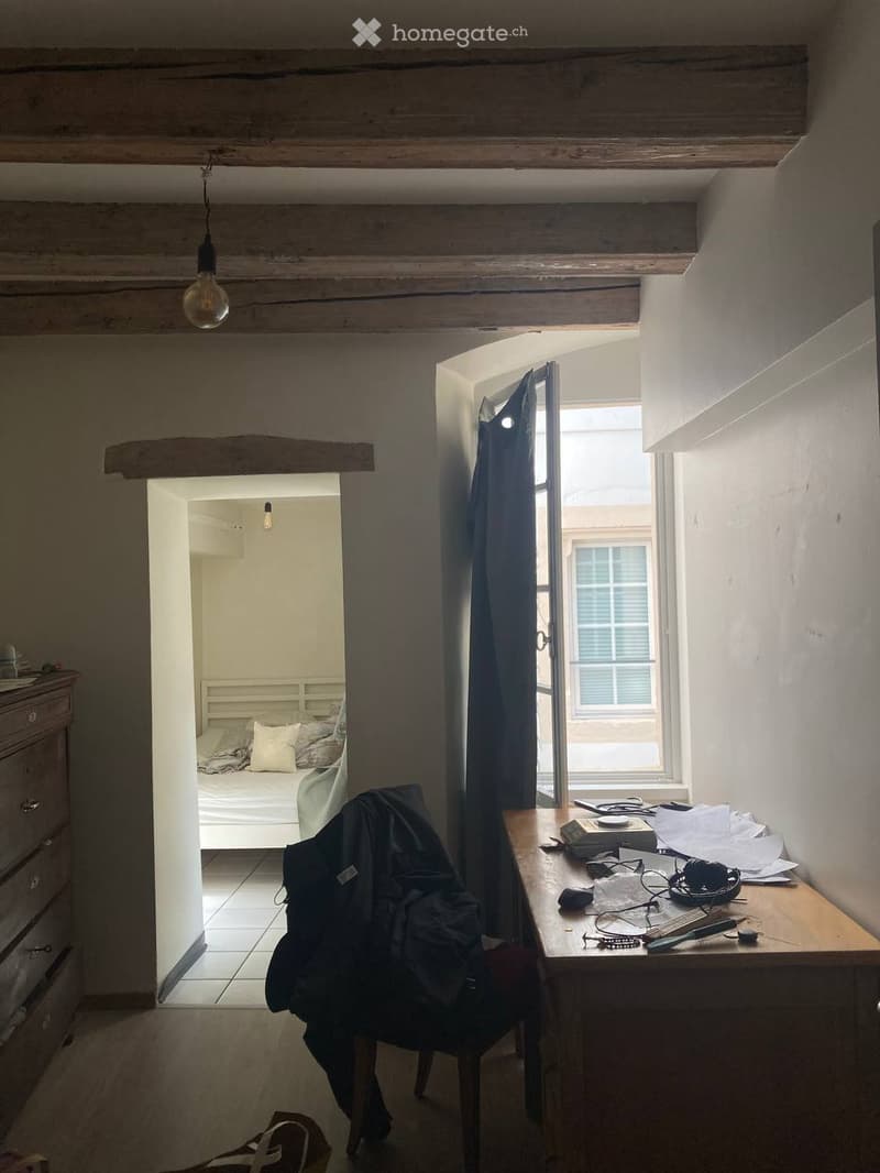 A small, rustic room with wooden beams and a wooden floor. A chair with a jacket draped over it is positioned near a wooden table holding a laptop, headphones, and papers. An open doorway leads to a bedroom with a bed, pillows, and a dresser.