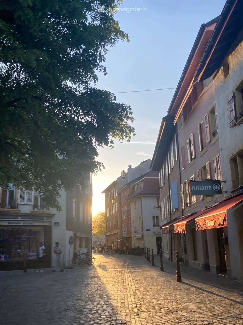 A picturesque cobblestone street lined with colorful historic buildings, featuring shops and cafes, with a tree providing shade and a warm sunset glow.