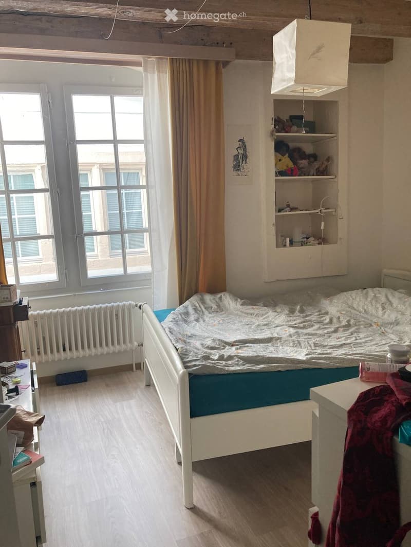 Cozy bedroom with a single bed, nightstand, radiator, built-in shelf, and large windows with curtains.