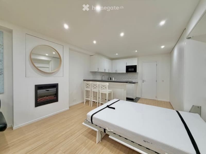 Modern, open-concept studio with a bed, fireplace, kitchen area, and seating area. Features include a round mirror, white stools, and sleek cabinetry.