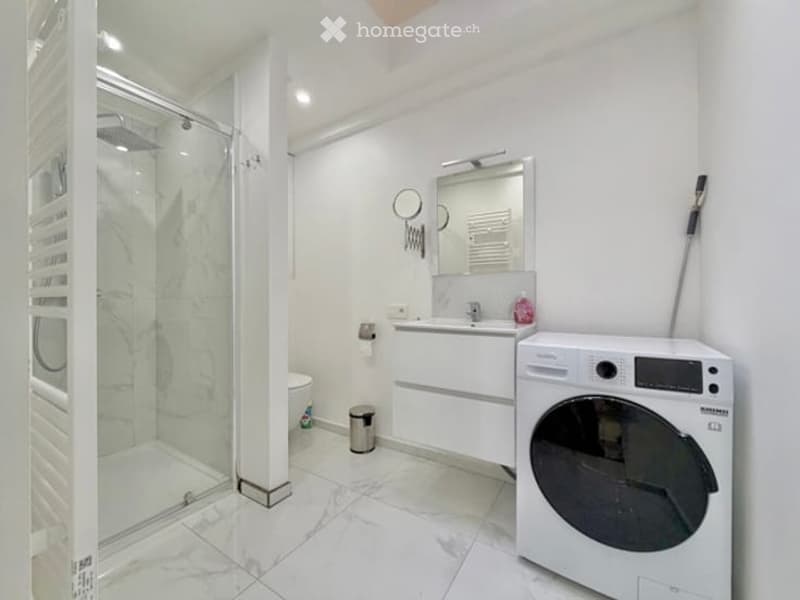 Modern bathroom with white tiles, a glass-enclosed shower, a sink with a mirror, a toilet, and a washing machine.