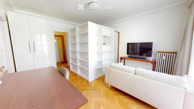 Bright, modern living room with white walls, wooden flooring, white sofa, coffee table, bookshelf, and TV on a stand.
