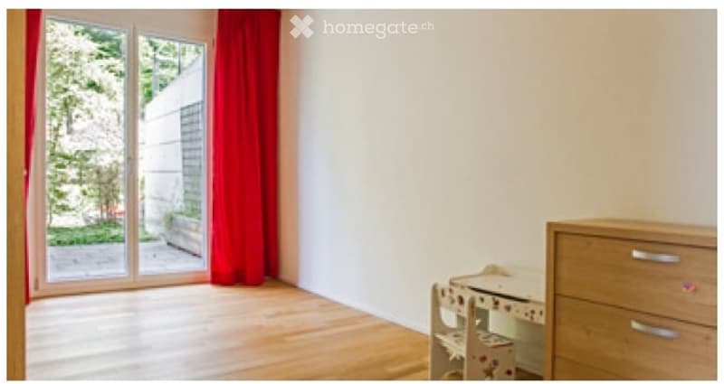 Bright, unfurnished room with wooden flooring, large sliding glass door leading to a garden, red curtain, and a small wooden dresser with a toy shelf.