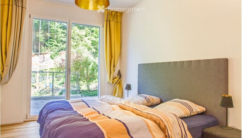 Modern bedroom with a large bed, headboard, pillows, and a view of greenery through large windows with yellow curtains.