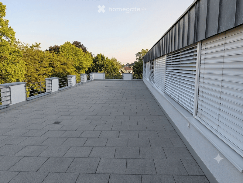 Modern balcony with grey tiled flooring, white railings, and white shutters on the adjacent building wall. The view includes green trees and a distant house.