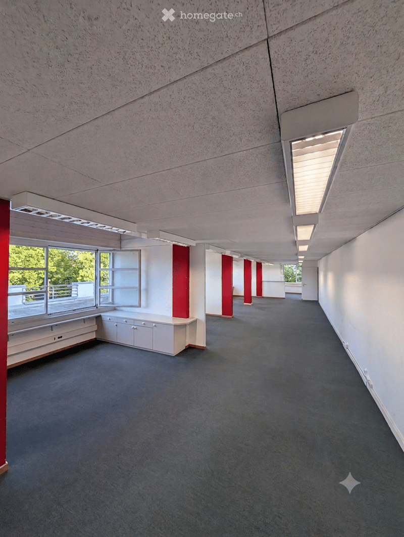 Empty hallway with grey carpet, white walls, red pillars, and large windows offering a view of the balcony area.