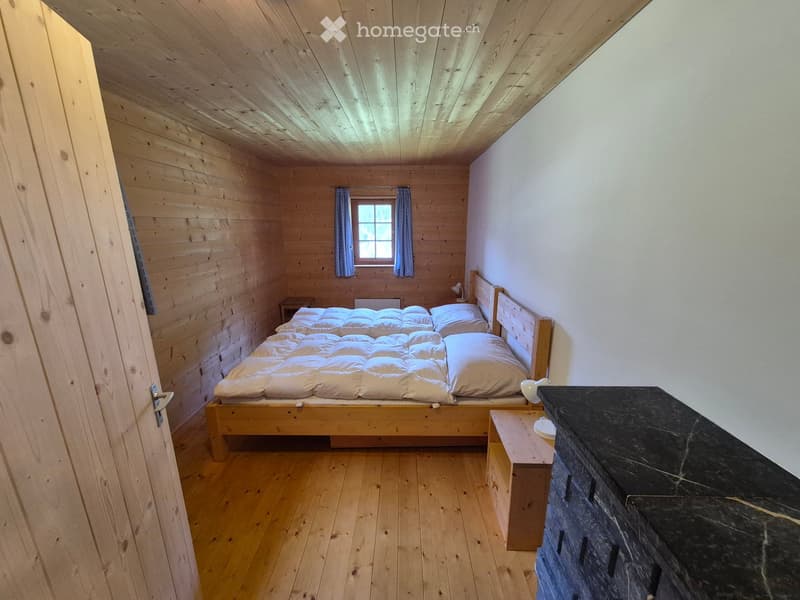 Cozy wooden bedroom with a neatly made bed, nightstand, and lamp. The room features wooden walls, ceiling, and floor, with a small window and a partially open door leading to another space.