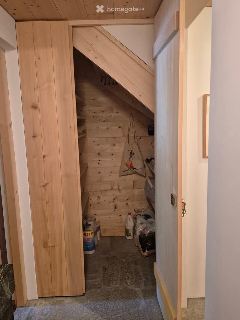 A narrow wooden staircase leading up, with storage shelves on the side and a few items stored, including a bag and cleaning supplies.