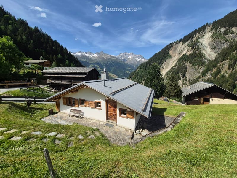 A small white wooden house with a grey metal roof, wooden shutters, and a wooden door, situated on a grassy slope with a stone pathway leading to the entrance. The surrounding area features other wooden houses, lush greenery, and distant mountain peaks.