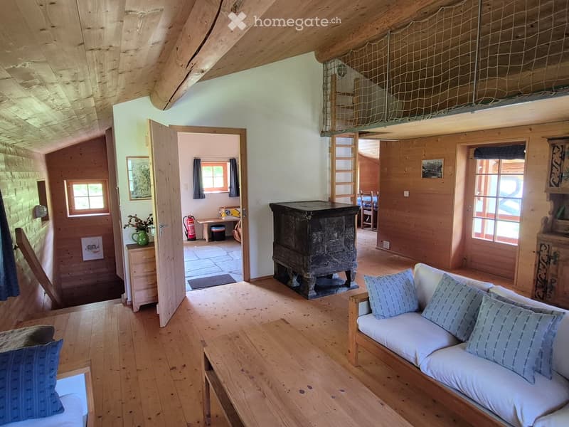 Cozy living room with exposed wooden beams, a white sofa with blue pillows, a wooden coffee table, and a large wooden cabinet. The room features wooden floors, large windows, and a view of another room with a fireplace and additional furniture.