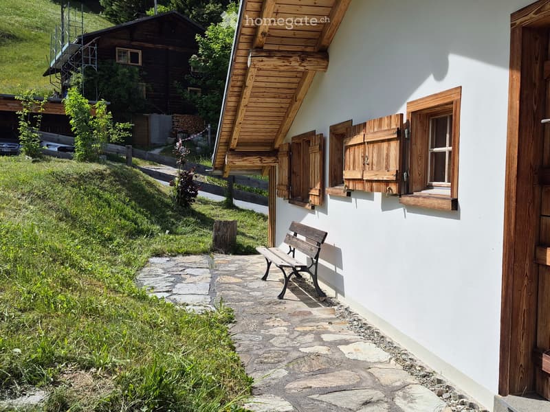 A white wooden house with a slanted roof and wooden shutters, featuring a stone pathway leading to a wooden bench, surrounded by greenery and a wooden fence.