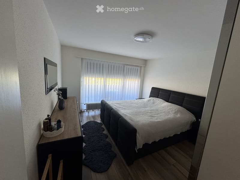 Cozy bedroom with a large bed, nightstand, mirror, vase, and rug. The room features wooden flooring and large windows with sheer curtains.