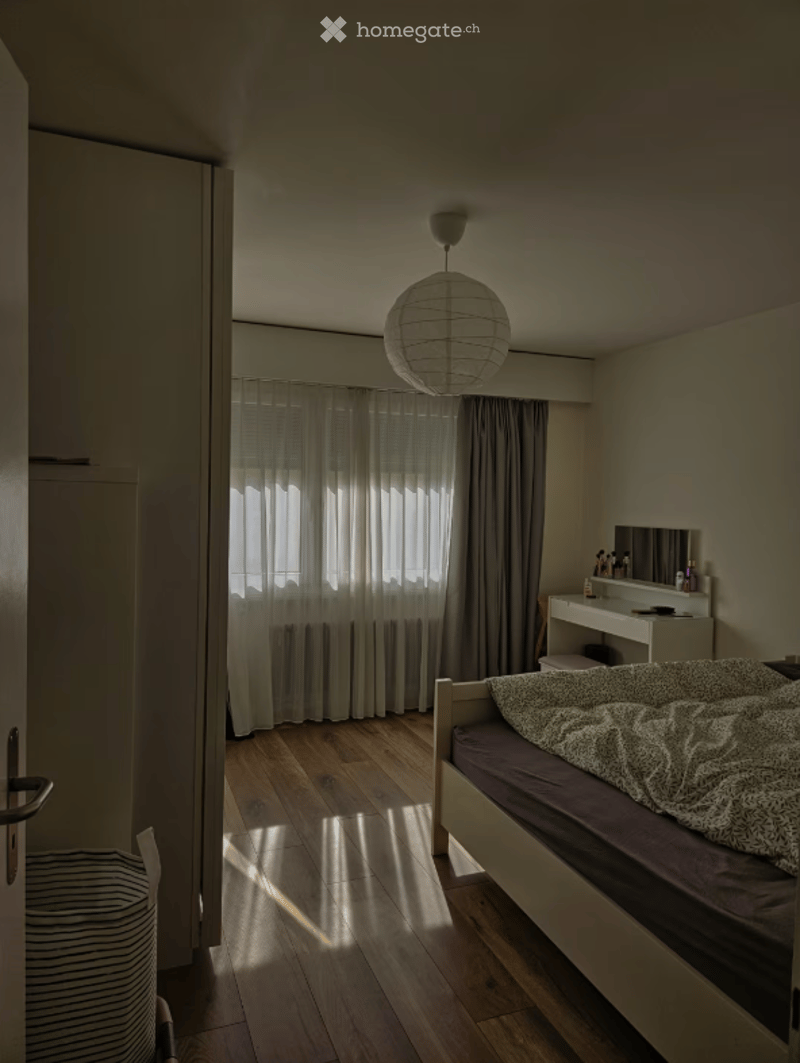 Bedroom. A modern, minimalist bedroom with a wooden floor, a single bed with a patterned duvet, a vanity with a mirror, and a hanging spherical lamp. The room features large windows with sheer curtains allowing natural light to filter in.
