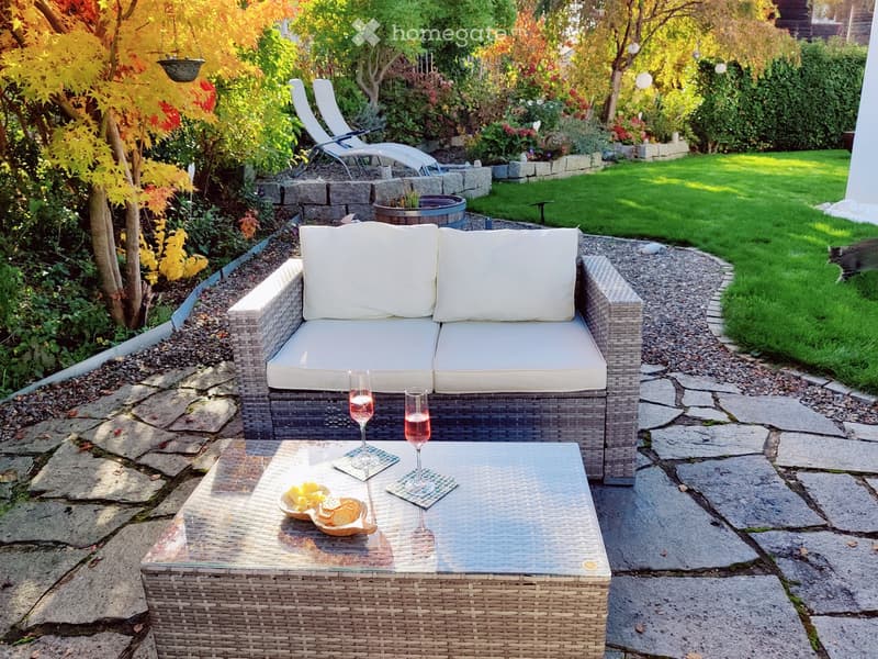 A well-maintained garden with a stone pathway, a wicker sofa with white cushions, a glass-topped coffee table, and two wine glasses with snacks.