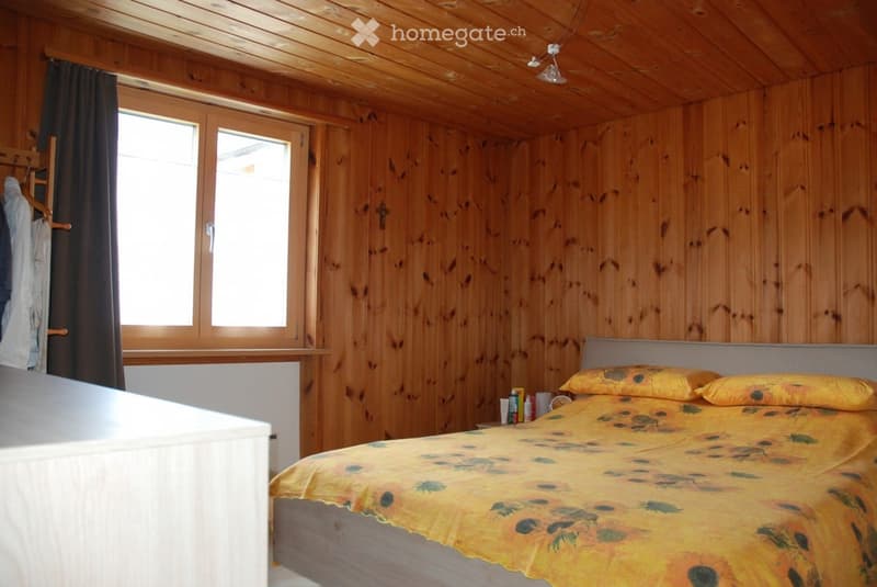 Cozy wooden bedroom with a bed, nightstand, and a window with curtains. The room features wooden walls and ceiling, and a small nightstand with a cup and books.
