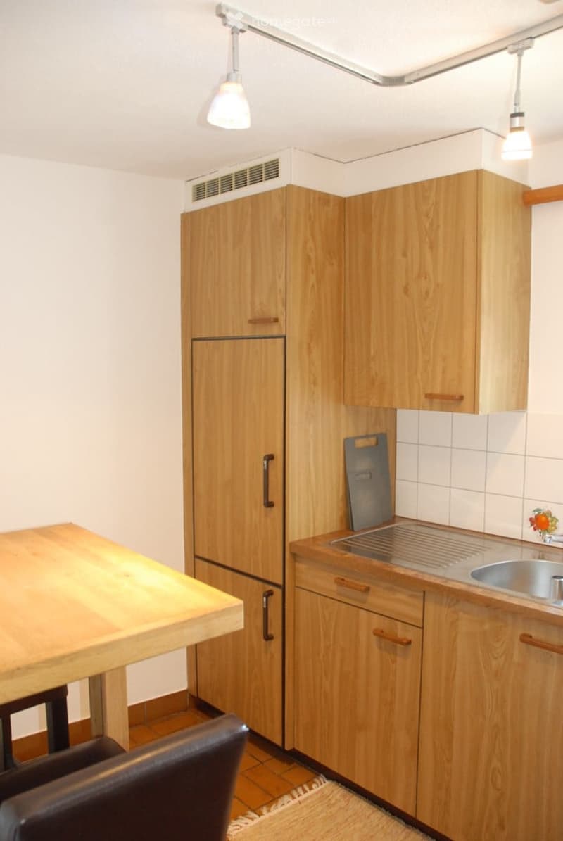Modern kitchen with light wooden cabinets, stainless steel sink, refrigerator, and a small wooden table with a chair.