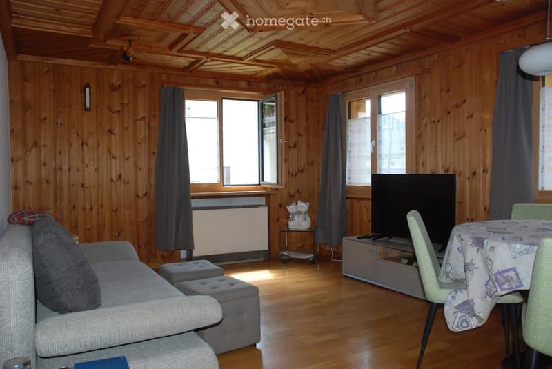 Cozy living room with wooden walls and ceiling, featuring a grey sofa with ottoman, a dining table with chairs, a television, and decorative items on a small table.