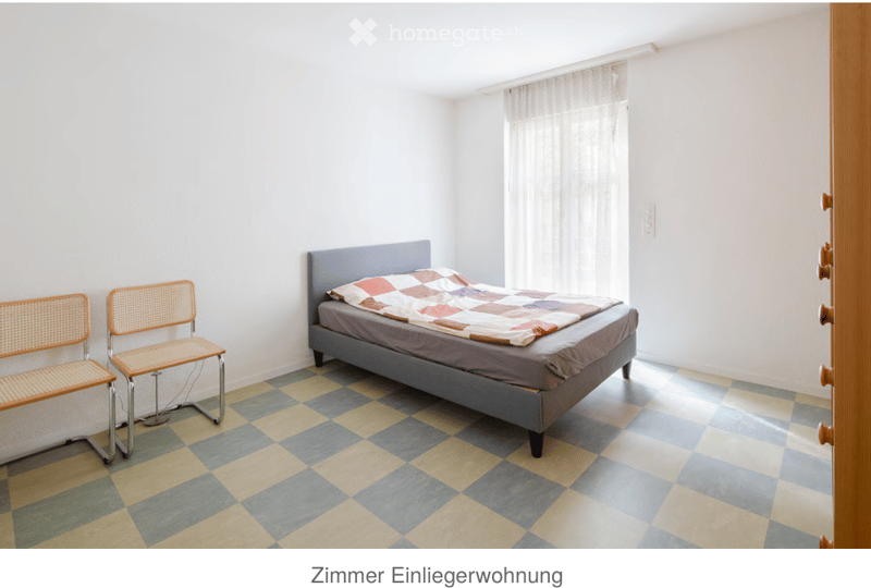 Bedroom with a single bed, two chairs, and a window with white curtains. The floor is checkered with blue and yellow tiles.
