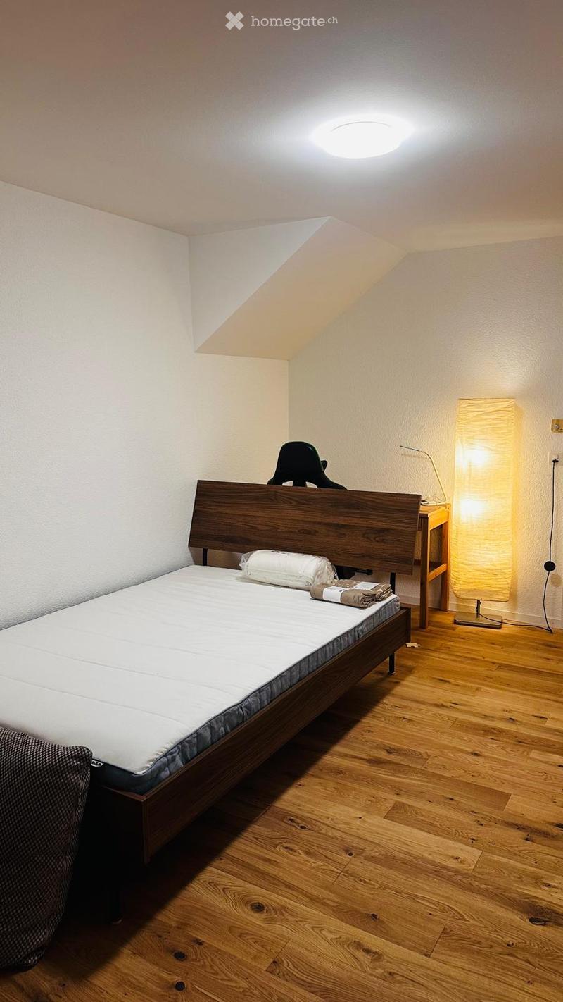 Cozy bedroom with wooden floor, a neatly made bed, a nightstand with a lamp, and a chair near the bed.