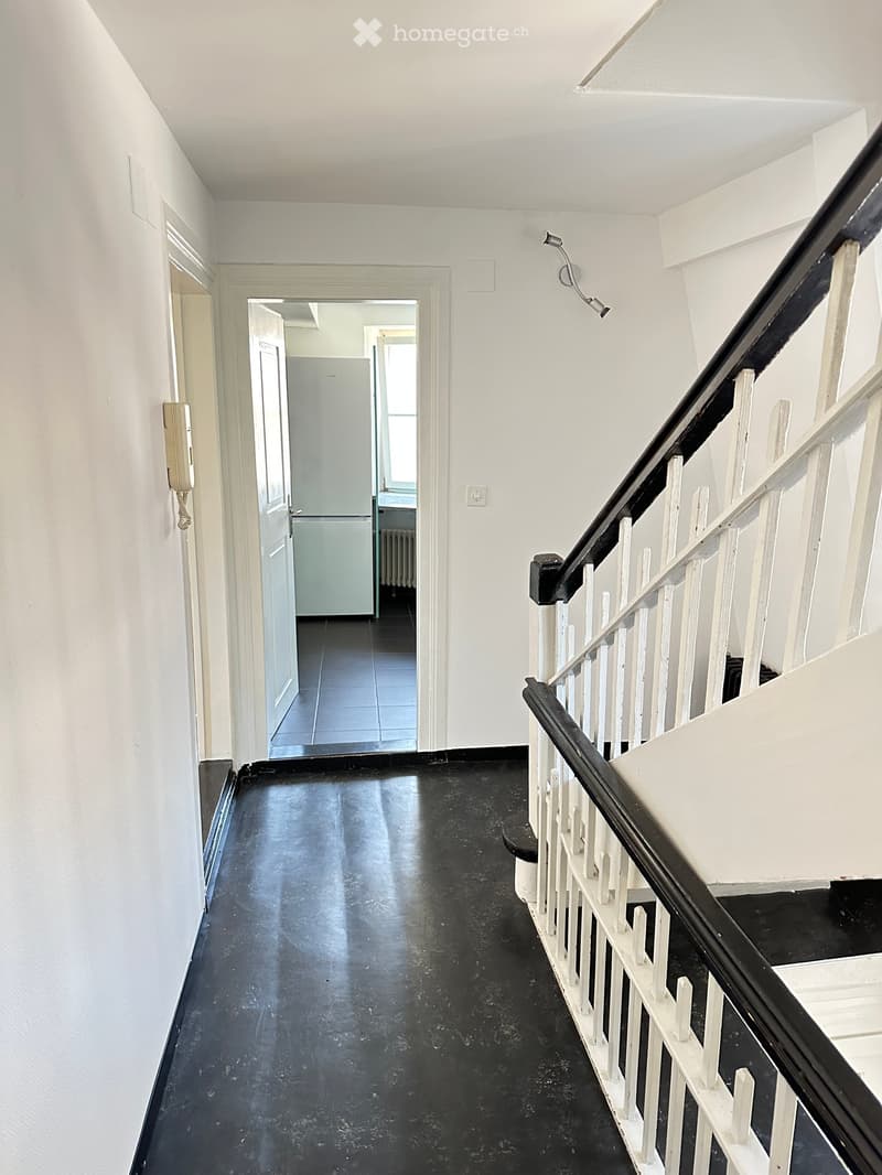 Empty hallway with black flooring, white walls, and a staircase with white railings. A door leads to a room with a refrigerator visible in the background.