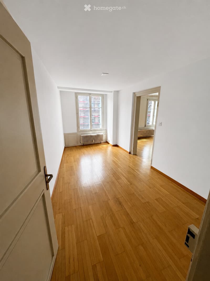Empty room with wooden flooring, white walls, and a window with a radiator below it. The room is connected to another space through an open doorway.