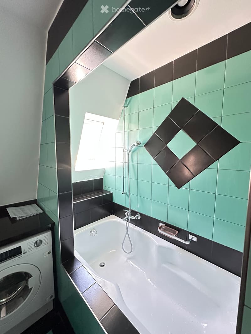 Modern bathroom with a bathtub, shower, washing machine, and a large mirror. The walls are tiled in green and black, and there is a window providing natural light.