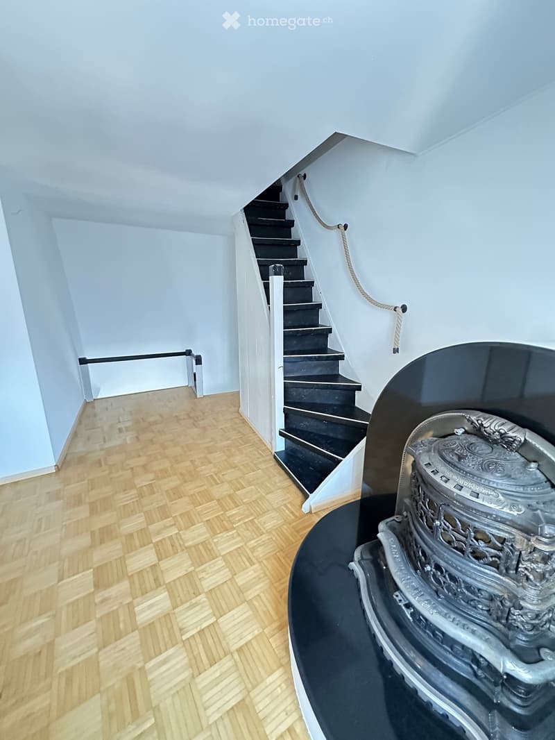 Spacious hallway with wooden herringbone flooring, white walls, and a modern black staircase with a rope handrail. A decorative ornate object is visible on a black platform to the right.