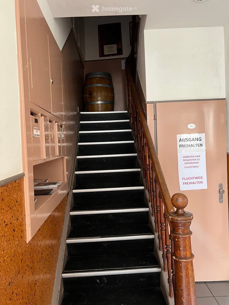A narrow staircase with black steps and a wooden handrail, featuring a barrel placed on the landing and a sign on the wall.