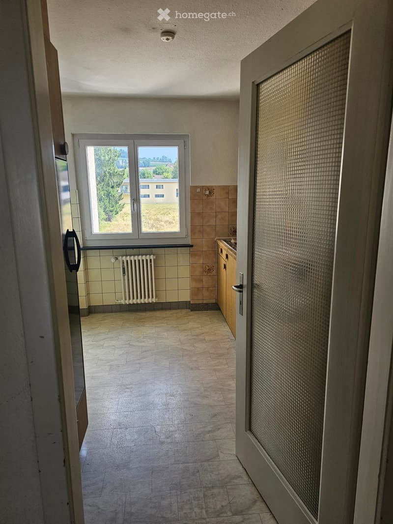 Empty room with tiled floor, beige walls, and a window with a view of the outdoors. There is a radiator below the window and a partially visible kitchen area with cabinets on the right side.