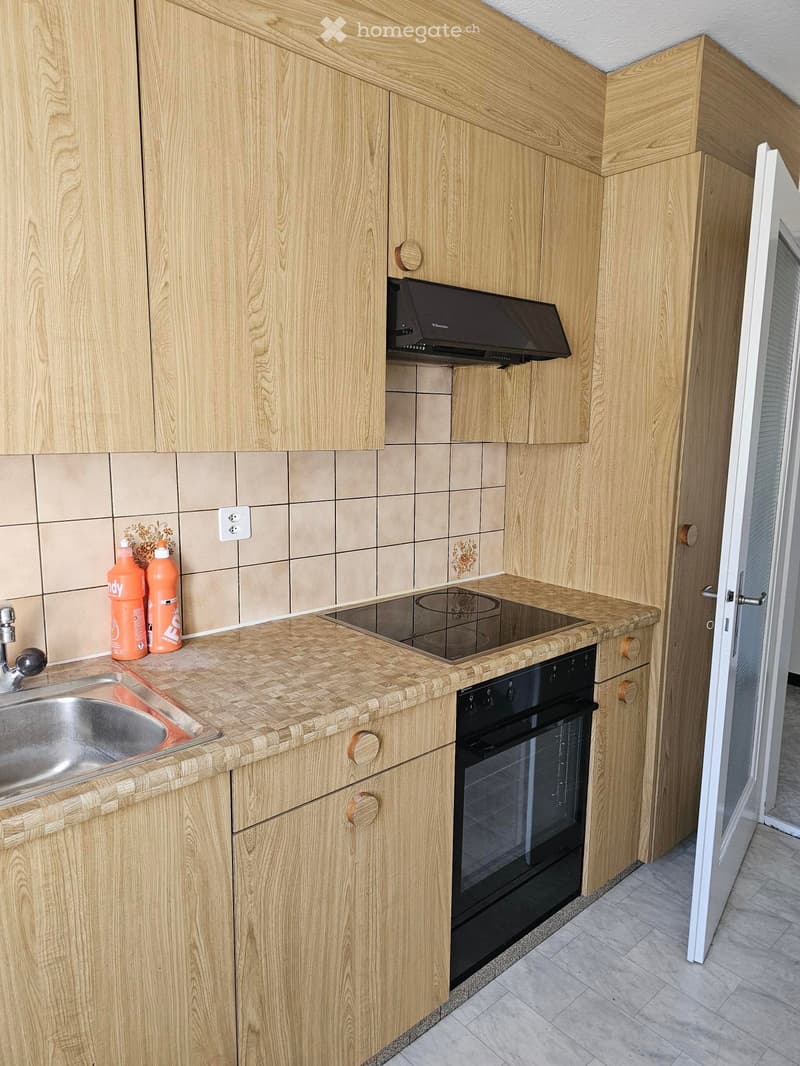Modern kitchen with light wooden cabinets, a stainless steel sink, an induction cooktop, and an oven. The countertop is made of a woven material, and there are two orange bottles on the counter.
