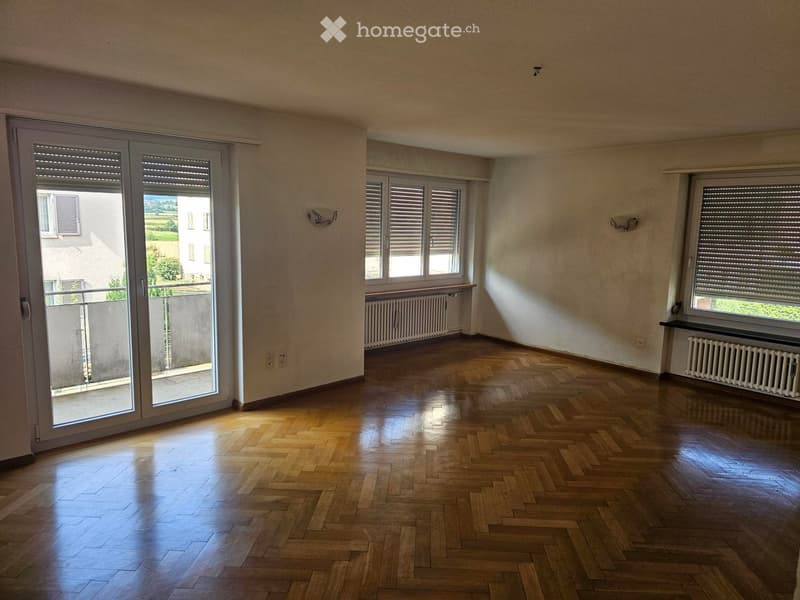 Empty room with wooden herringbone flooring, large sliding glass door leading to a balcony, and multiple windows with blinds.