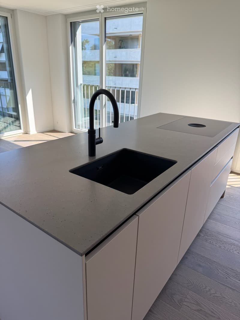 Modern kitchen with a sleek, grey countertop, a black sink, and a contemporary black faucet. The kitchen features light-colored cabinetry and large windows with a view of the balcony.