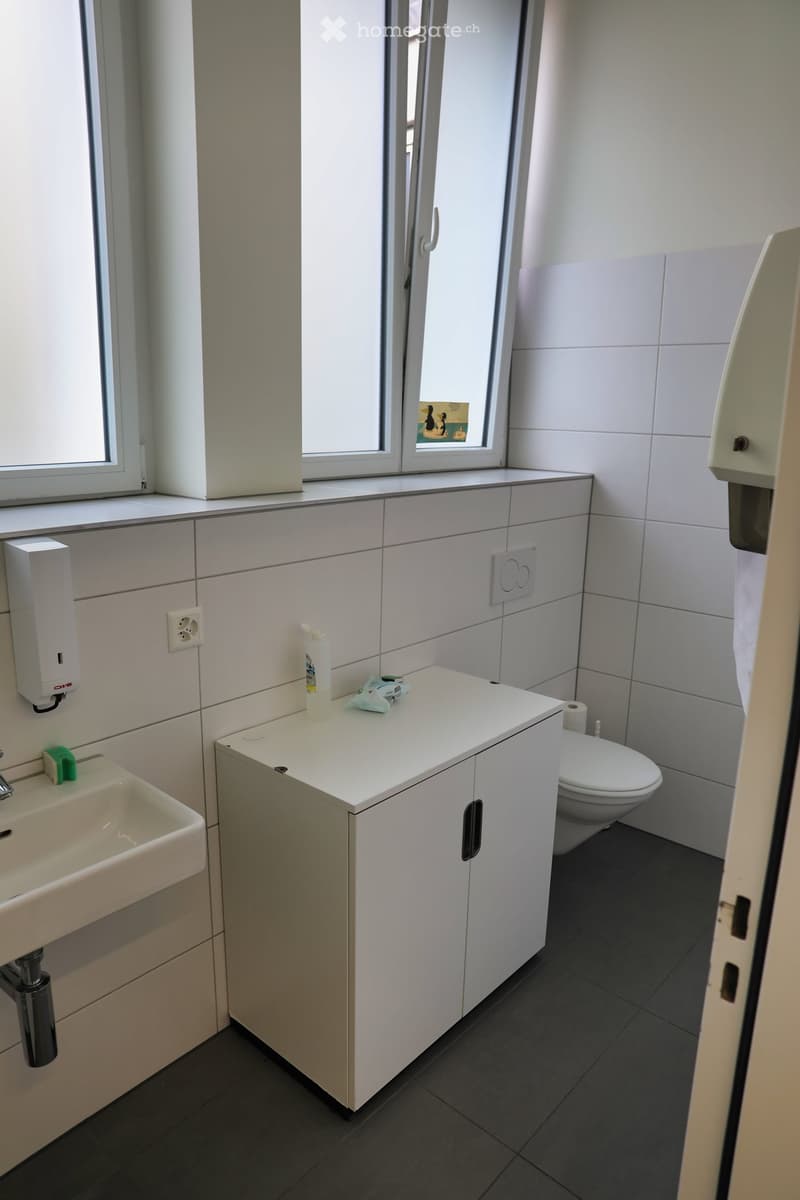 white tiled walls, white sink, white toilet, small cabinet with white door, white hand soap dispenser, black floor tiles