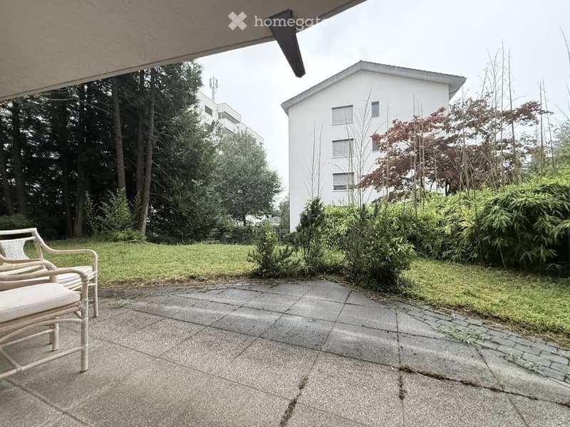 Garden area with a paved patio, two wicker chairs, and surrounding greenery including bushes and trees.