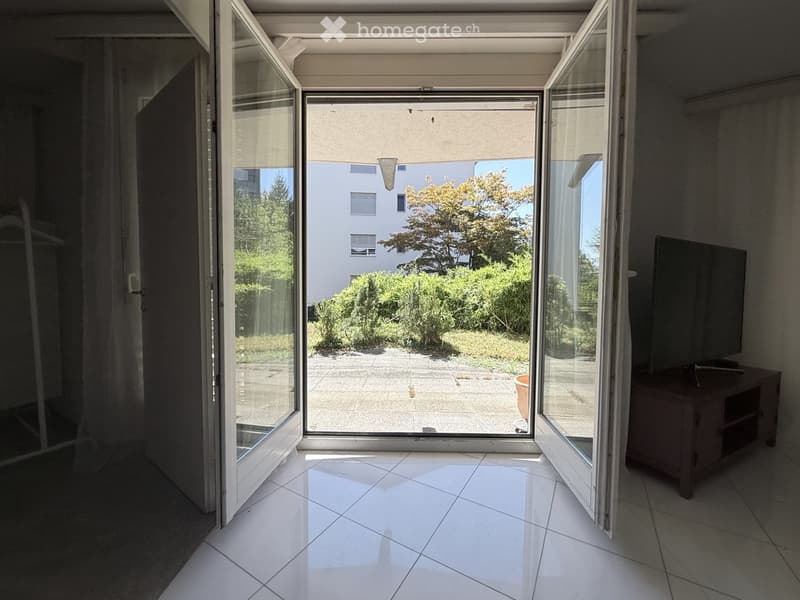 Bright room with large sliding glass doors opening to a patio, tiled floor, TV on a stand, and a view of greenery and neighboring building.