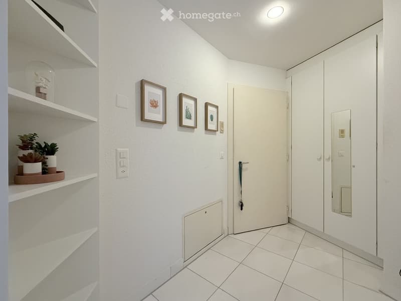 A narrow hallway with white tiled flooring, white walls, built-in shelves on one side, and framed artwork on the wall. A closed door is visible at the end of the hallway.