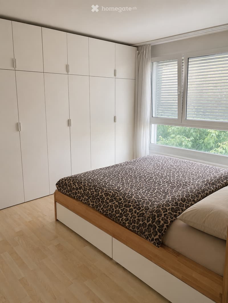Modern bedroom with a wooden bed, leopard print duvet, white wardrobe, and large windows with blinds.