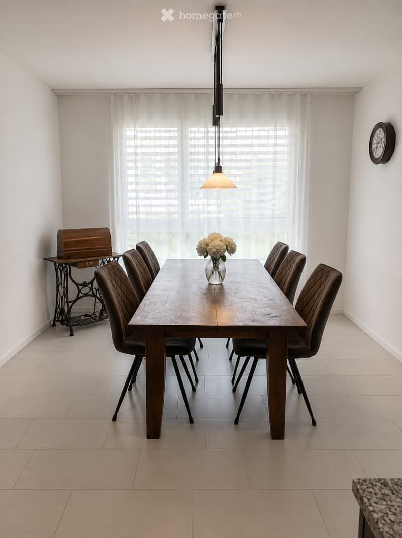Dining room with a wooden dining table, six brown upholstered chairs, a vase with white flowers, a small wooden stool, and a wall clock.