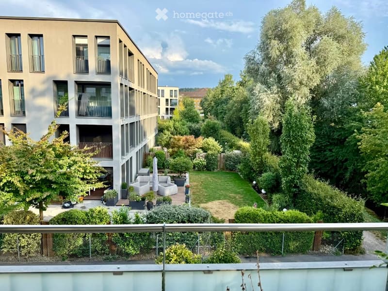 A well-maintained garden area with a lawn, various trees, shrubs, and outdoor seating including a sofa and chairs. The garden is bordered by a modern building with balconies and large windows.